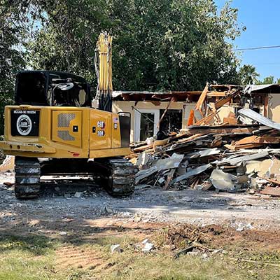 A yellow excavator demolishing a house, surrounded by debris and tree in the background.