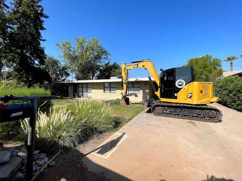 A yellow excavator parked on a concrete driveway in front of a single-story house with a grassy yard and trees under a clear blue sky.