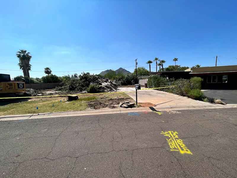 A vacant lot with scattered debris, palm trees, a distant mountain, and a construction vehicle under a clear blue sky. Yellow markings are visible on the pavement.