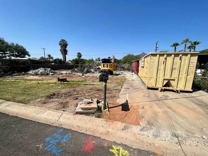 Construction site with cleared land, debris, a yellow dumpster, and equipment under a clear blue sky. Palm trees and caution tape are visible in the background.