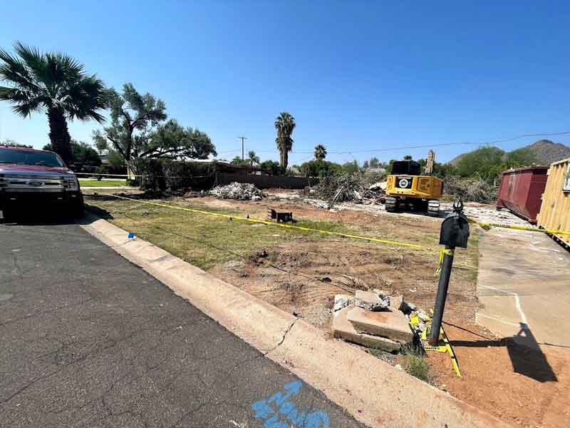 Construction site with rubble, a yellow excavator, a red dumpster, and caution tape on a clear day. Nearby are palm trees and a parked vehicle on the left.