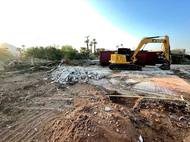 Yellow excavator on a construction site with debris and rubble in the foreground, trees and a clear sky in the background.