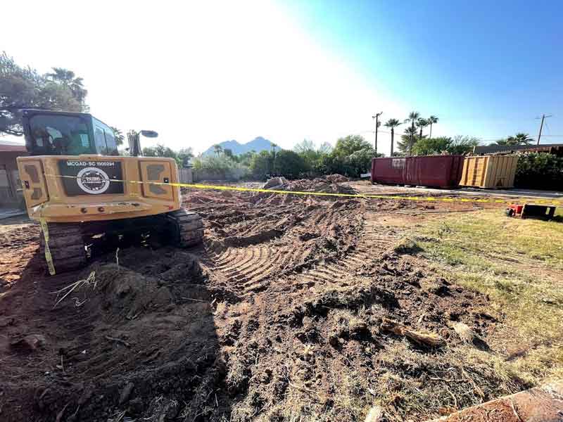 Construction site with bulldozer, soil, and yellow tape. Shipping containers in the background and trees lining the area.