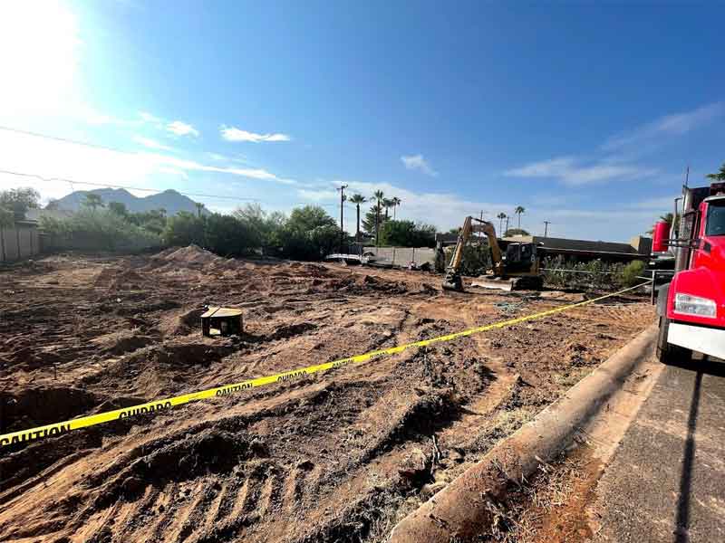 Construction site with excavator and caution tape on dirt ground under a clear blue sky, surrounded by trees and utility poles.