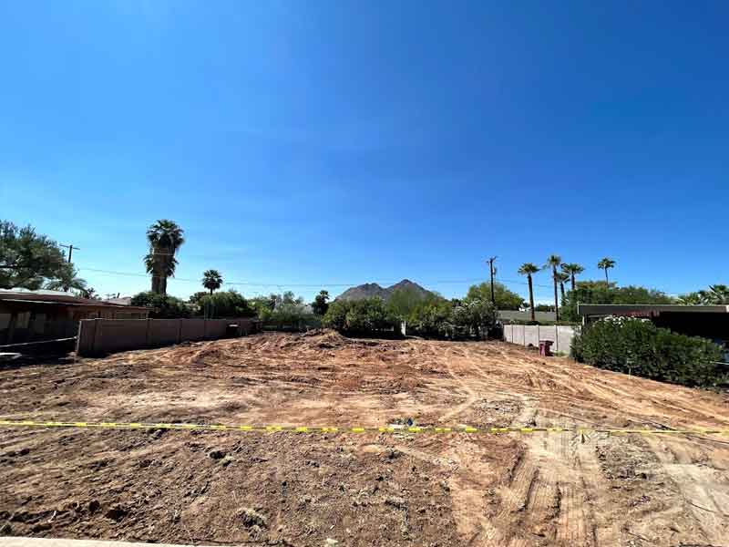 Cleared plot of land with a dirt surface, surrounded by trees and bushes, mountain visible in the background under a clear blue sky.