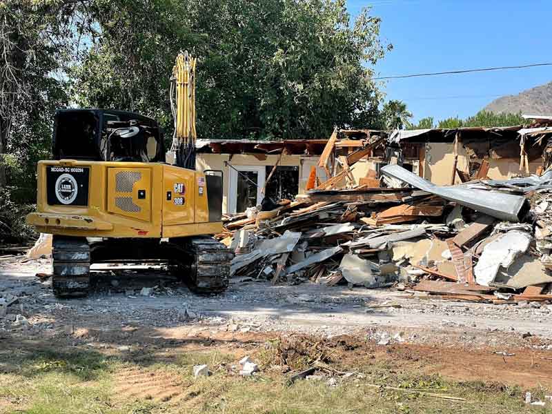 A yellow excavator demolishing a partially collapsed house with debris scattered in front. Trees and blue sky in the background.