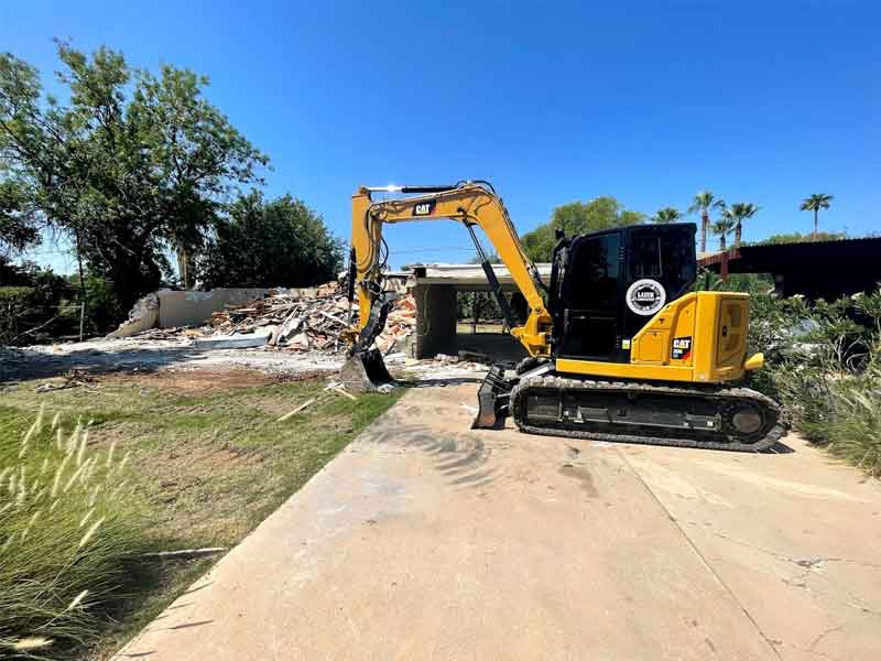 A yellow excavator is parked on a concrete path next to a pile of debris from a demolished building, with trees in the background under a clear blue sky.