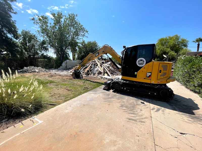 A yellow excavator sits on a driveway near a pile of debris and rubble, surrounded by trees.