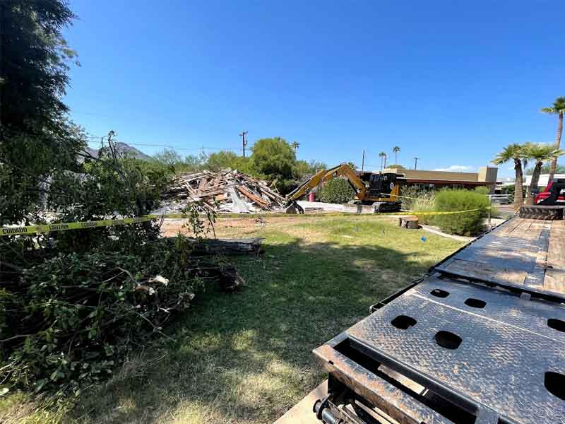 A construction site with a pile of debris and an excavator. Caution tape is visible, along with trees and a clear blue sky.