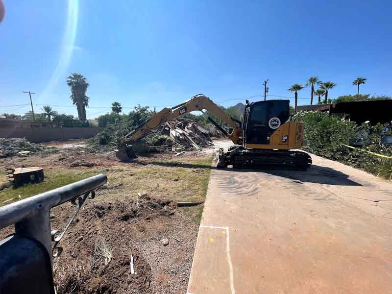 A yellow excavator clears debris in a yard with palm trees in the background under a clear blue sky.