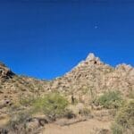 Rocky desert landscape with cacti under a clear blue sky and a small white moon visible.