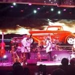 Band performing on stage with a large screen displaying a red vintage car in the background. Musical instruments are visible on stage.