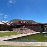 The Taliesin West building with angled red beams, stone walls, and a reflective pool in front. Grass surrounds the area, set against a mountain and blue sky backdrop.