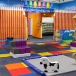 A colorful children's play area featuring crayon-themed decor, cushioned seats, and playful animal ride-on toys on a patterned floor.