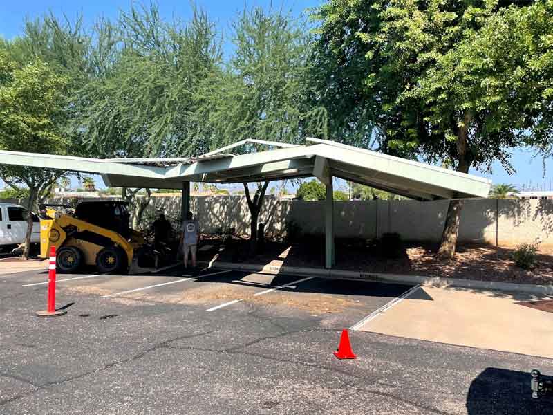 A damaged carport leaned over in a parking lot, supported by a utility vehicle, with traffic cones marking the area.