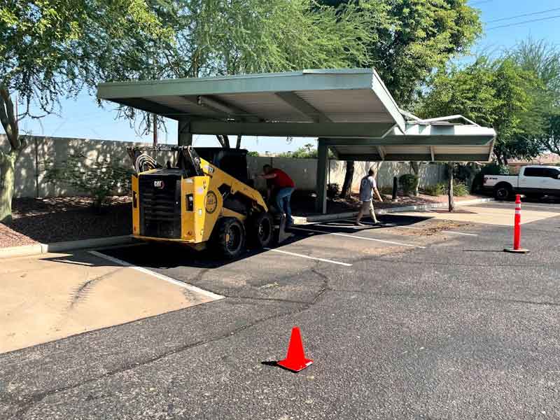 A small construction vehicle and two people work under a metal carport in a parking lot. Traffic cones are placed nearby for safety.