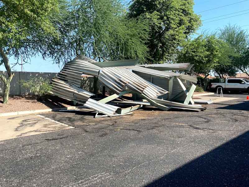 A collapsed metal structure lies on an asphalt surface near trees and a parked vehicle.