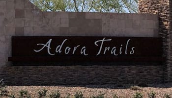 Sign reading "Adora Trails" on a stone wall with desert landscaping in the foreground.