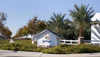 Entrance sign surrounded by shrubs and trees near a white fence under a clear sky.