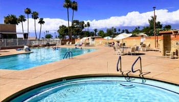 Outdoor swimming pool area with a circular hot tub in the foreground, lounge chairs, tables with umbrellas, and palm trees under a clear blue sky.