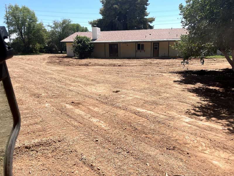 A house with a beige exterior stands surrounded by a large, bare dirt yard and sparse trees under a clear sky.