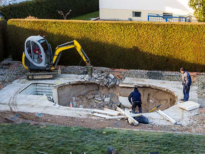 A construction site with a small excavator and two workers removing debris from a partially demolished swimming pool.