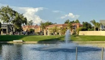 A serene pond with a central fountain is surrounded by greenery and buildings under a clear blue sky.