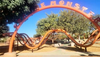 Orange metal playground structure resembling a dragon with wavy slides and climbing areas, set in a park with trees.