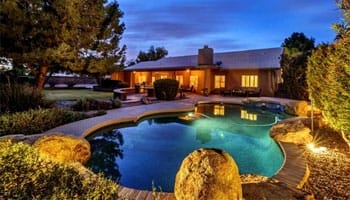 Backyard with a kidney-shaped pool surrounded by rocks and landscaping, adjacent to a single-story house under a twilight sky.