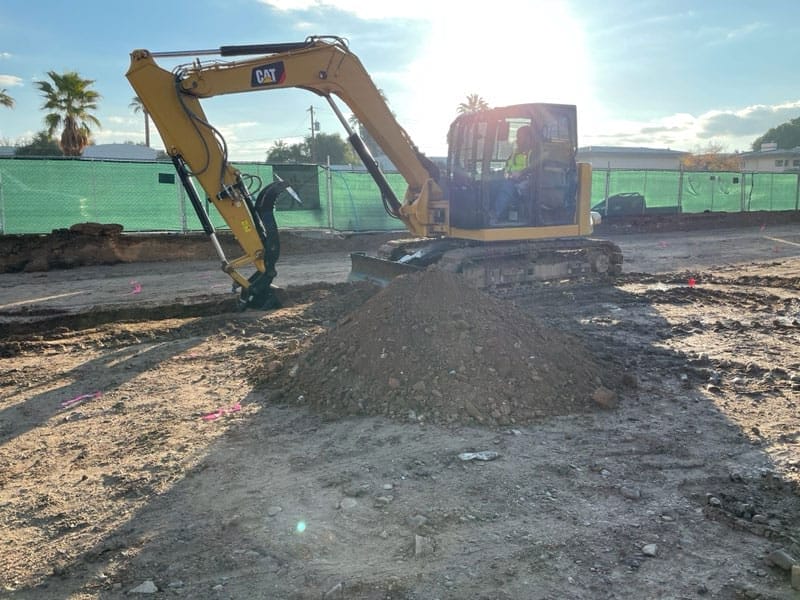 Yellow excavator moving dirt on a construction site with a green fence and palm trees in the background.