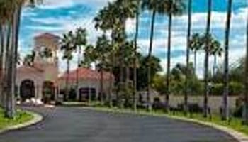 A curved driveway lined with tall palm trees leads to a large building with a red-tiled roof under a partly cloudy sky.