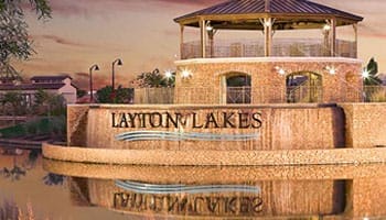 A brick pavilion with "Layton Lakes" sign, next to a reflective body of water, under a sunset sky.