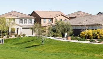 A row of suburban houses with beige and brown tones, surrounded by green lawns and small trees under a clear sky.