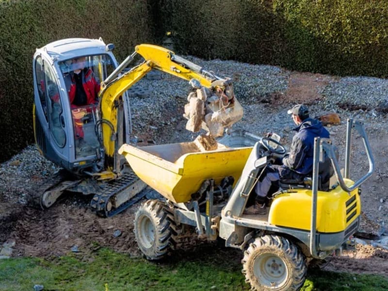 A small excavator loads dirt from a partial pool fill project in into a yellow dumper truck in a fenced yard in Mesa AZ. Two workers are operating the machinery.