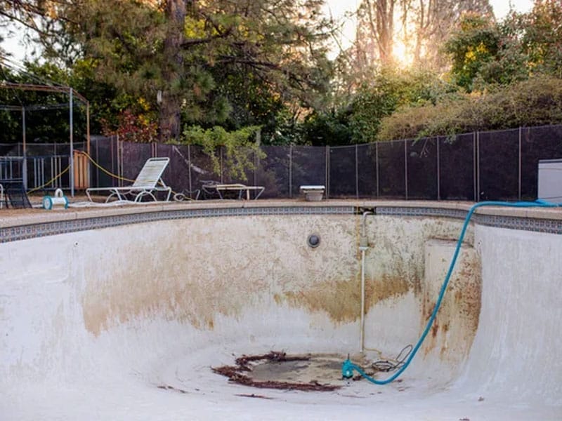 An empty, dirty swimming pool with a hose inside, surrounded by trees and outdoor furniture while we wait for demo permits from the city of Mesa AZ.