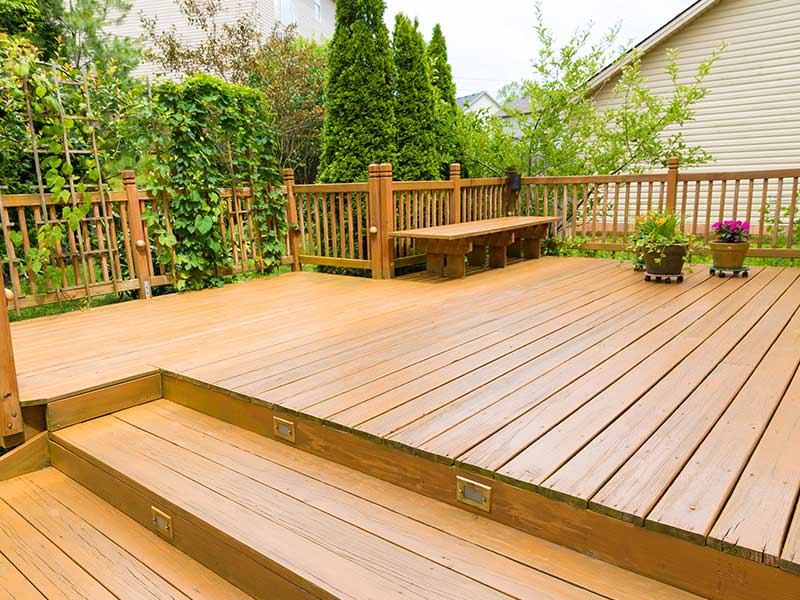 A wooden deck with stairs built over a filled-in swimming pool, surrounded by a fence and greenery. Potted plants are placed along the wall, and a wooden bench is visible on one side.