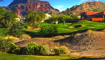 A lush golf course set against a backdrop of red rocky hills and clear blue sky, with a modern building nearby.