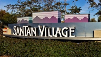 Sign for SanTan Village, featuring a stylized mountain range in the background. Surrounded by greenery and trees under a blue sky.