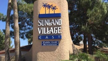 Sign reading "Sunland Village East" with icons of palm trees and a sun, surrounded by trees and greenery.