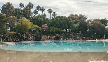 A tranquil outdoor swimming pool surrounded by trees and rocks under a cloudy sky.